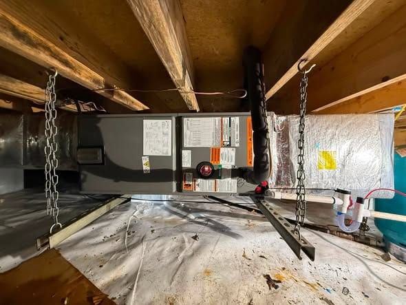 Air handler hung from chains between floor joists in a crawlspace, with its drain line and foil wrapped duct beside it