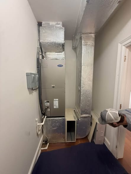 Indoor air handler in a narrow hallway closet with foil wrapped ducts above and beside it and a drain pipe at its base